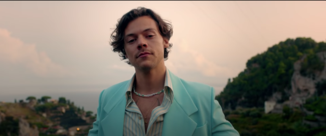Harry Styles smirks at the camera in a blue suit with a beaded necklace. Photo courtesy of YouTube.