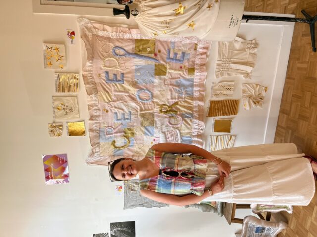 Standing in front of her quilted tapestry, Caitlynn Reynolds displays her final sustainable textile project. The phrase “Created to Create” highlights her belief in honoring handmade craft and storytelling through fabric.