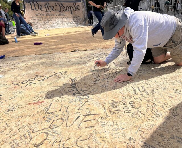 A man writes "Money for school, not for war," on the Constitution banner.